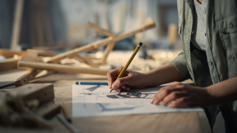 Close Up Portrait Of A Furniture Designer Writing Down Dimensions And Assembling Legs Of A Wooden Chair. Stylish Black Female Carpenter Working In Studio In Loft Space With Tools On The Walls.